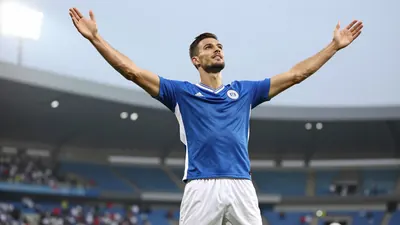A photorealistic image of a tall, muscular male footballer in a blue jersey and white shorts, celebrating a goal with...
