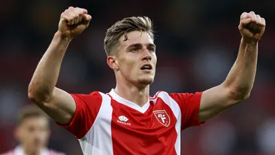 A 22-year-old Scottish striker in a red and white striped shirt, celebrating a goal with his arms raised