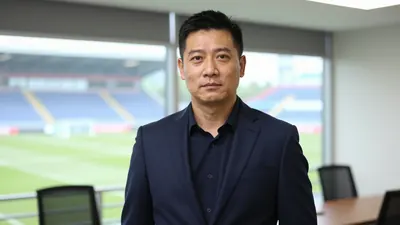 A 35-year-old male club director in a dark blue suit, standing in a modern office with a blurred view of a football...