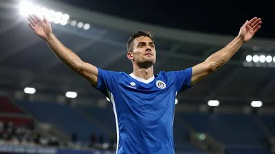 A 25-year-old male football player in a blue jersey celebrating a goal with his arms raised