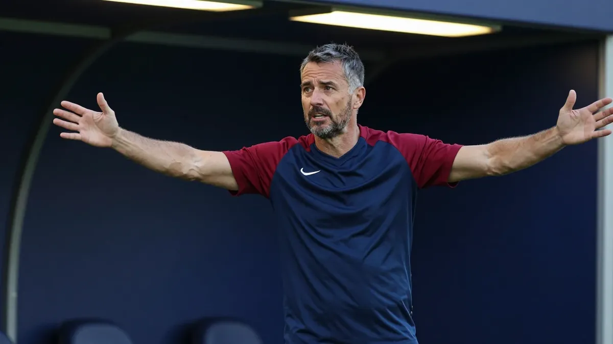 A 35-year-old, 5'10" assistant coach with a rugged build, dressed in a navy and claret plain, unmarked training top made...
